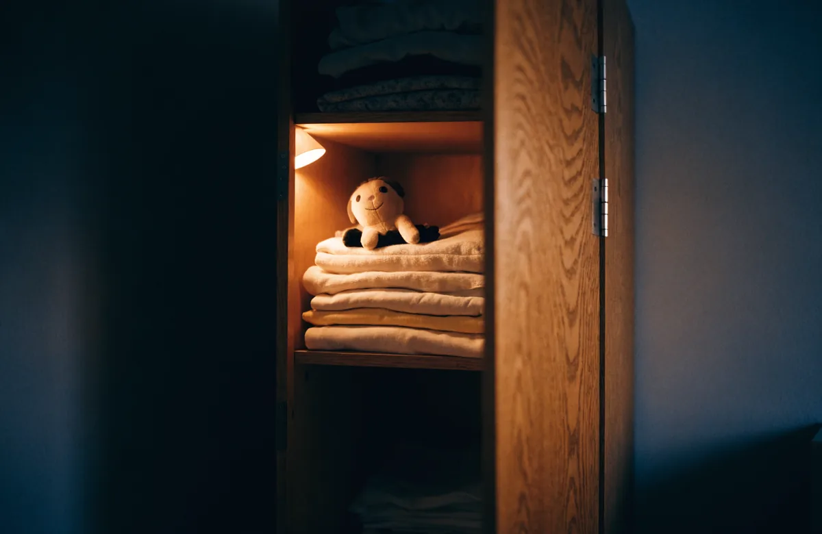A soft toy sitting on a pile of folded blankets inside a lit wardrobe with its door open.