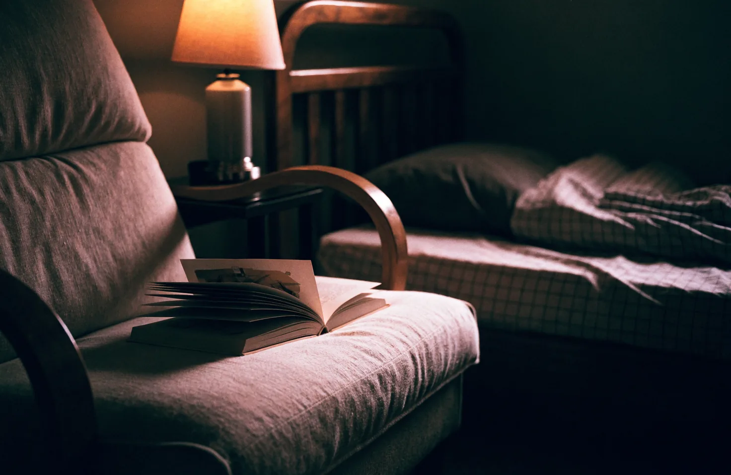 An open book resting on the arm of a nursing chair, beside a child’s bed and a lit lamp.