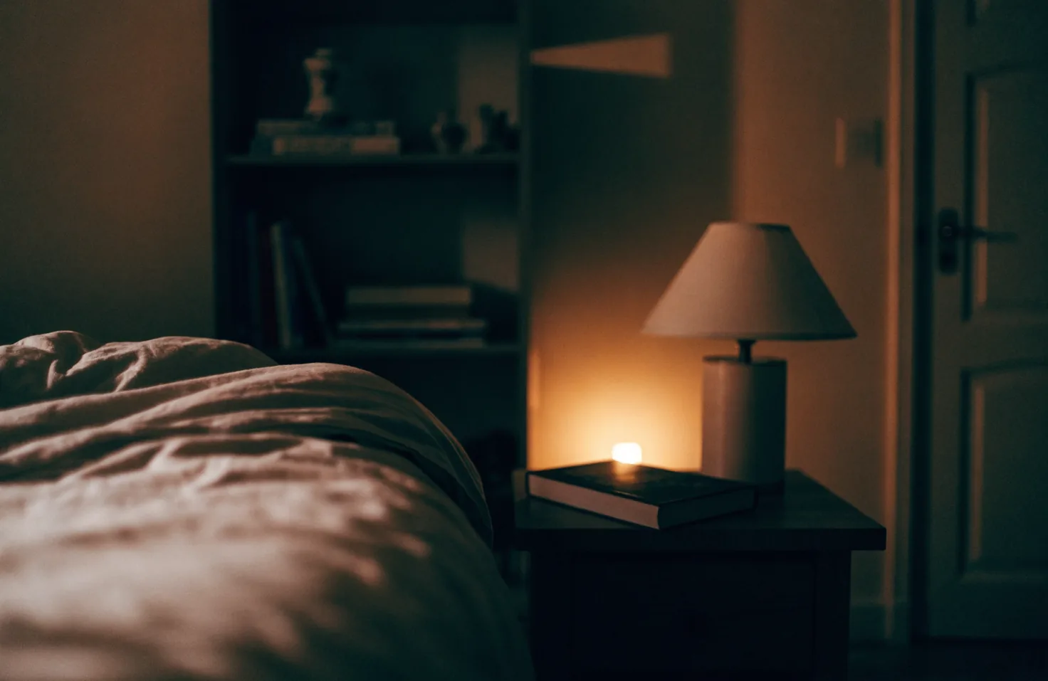 A lamp and a closed hardback on a bedside table, the bed in shadow and the bedroom door standing ajar.