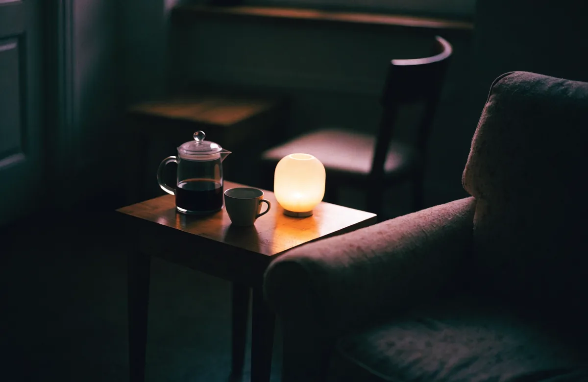 A glass coffee pot, a cup and a small lamp on a side table next to an armchair in an otherwise dark room.