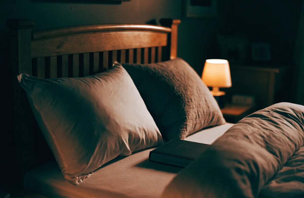 Two pillows against a wooden headboard and a turned down duvet, a closed notebook on the sheet, a lamp lit beyond.