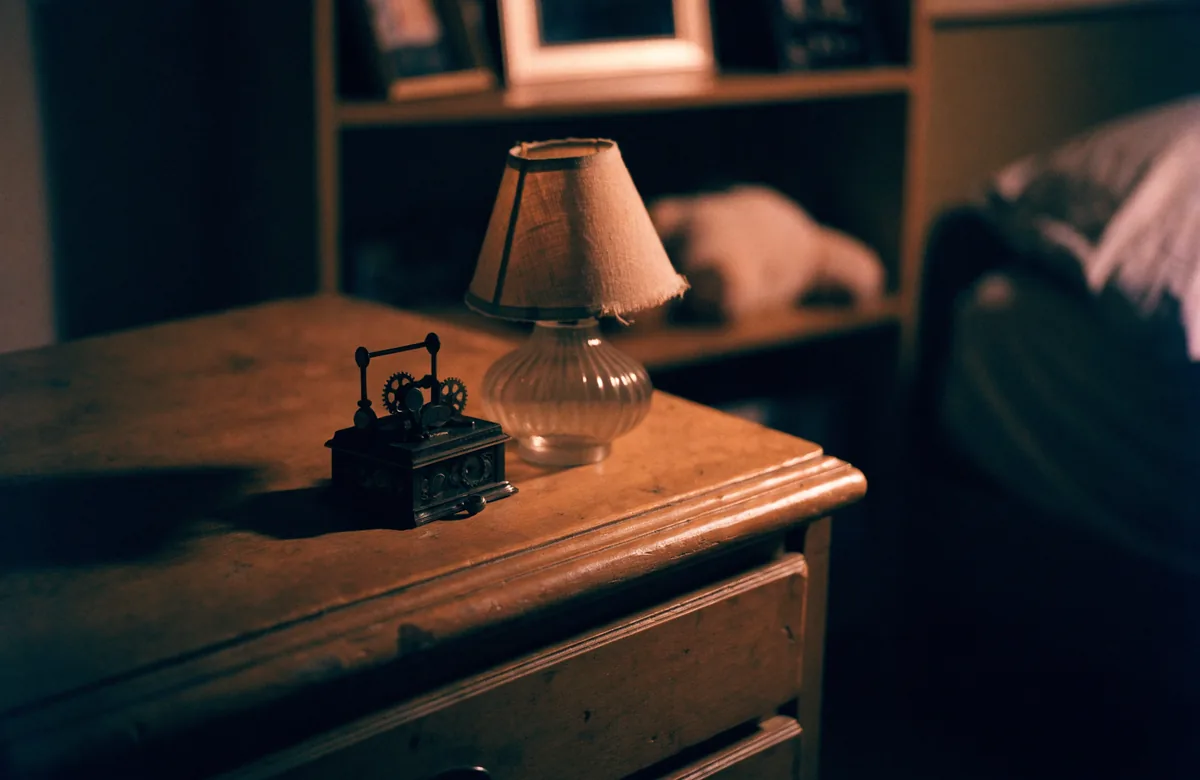 A wind up music box on a wooden chest of drawers next to a small lamp, a child’s bed behind it.